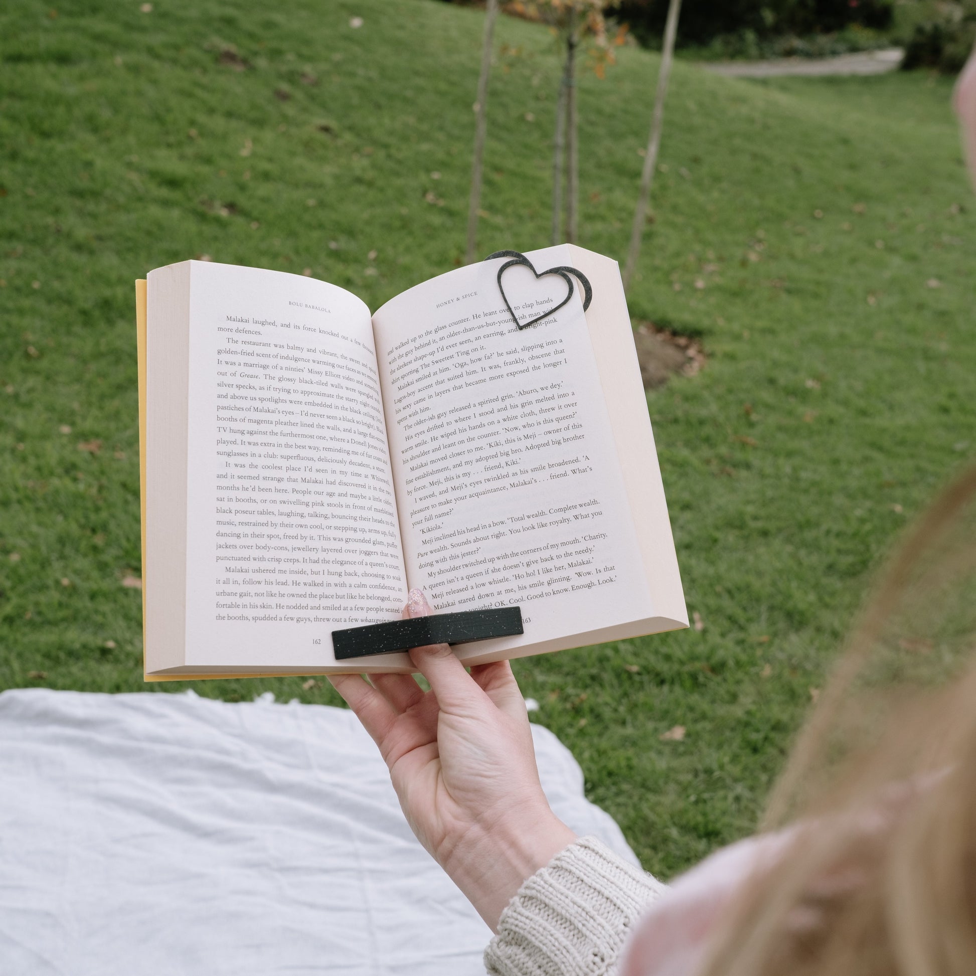 Person reading an open book with a heart bookmark on a grassy field